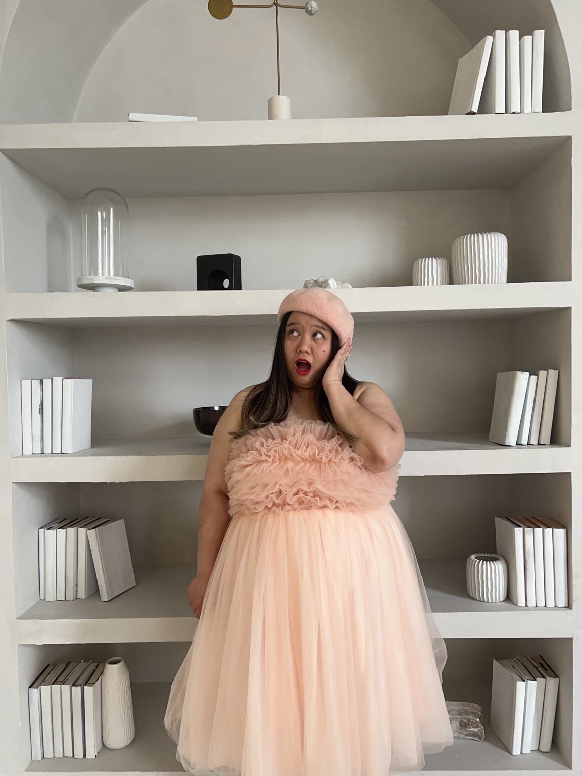 Dee in a peach dress and pink beret beside sculptural white shelves